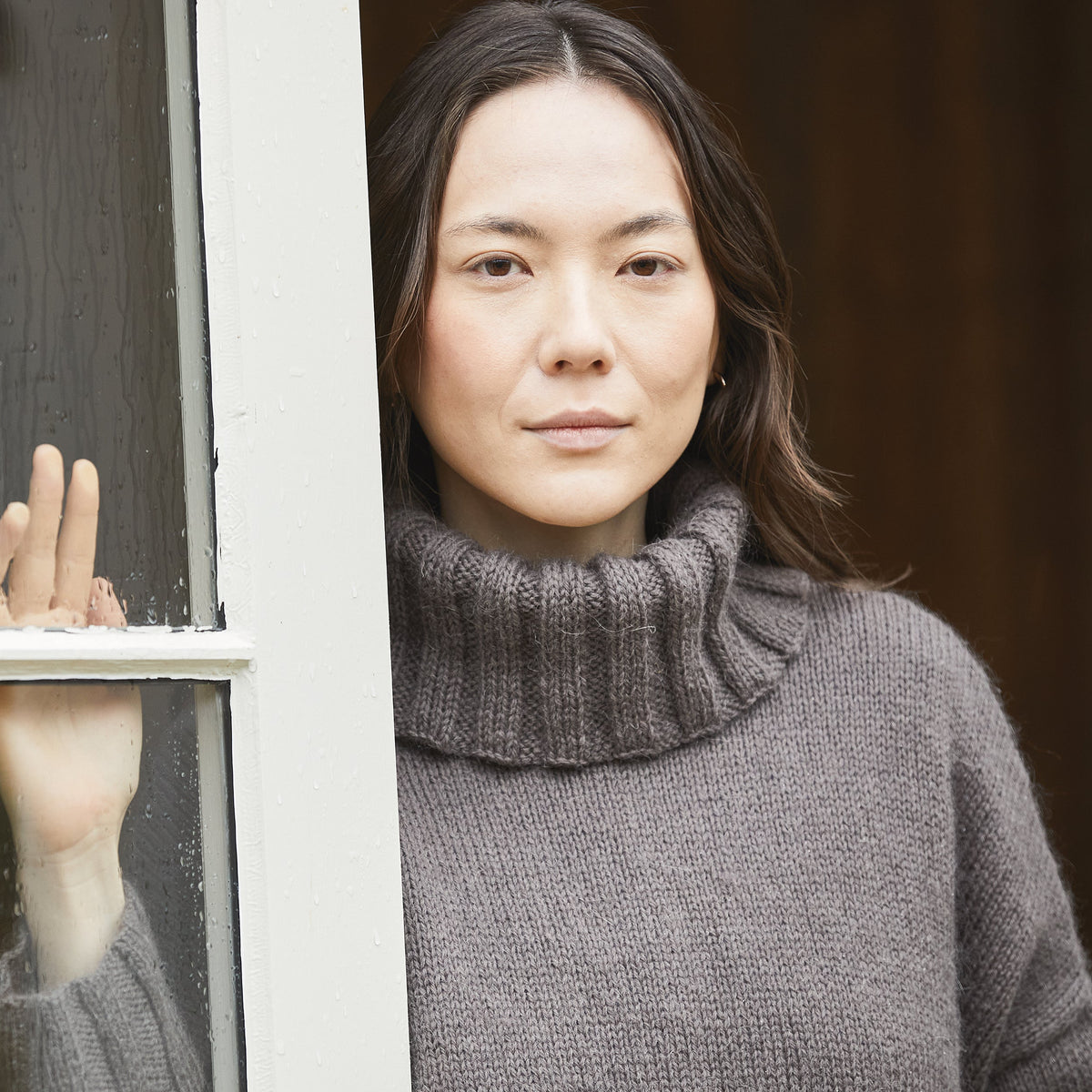Woman in a gray sweater standing by a window with raindrops on the glass.