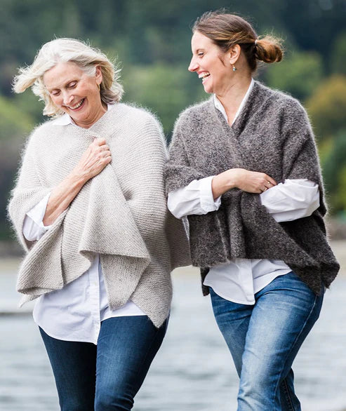 Two women standing by a body of water, both wearing gray quadrangle wraps with V-necklines, paired with white shirts and blue jeans.