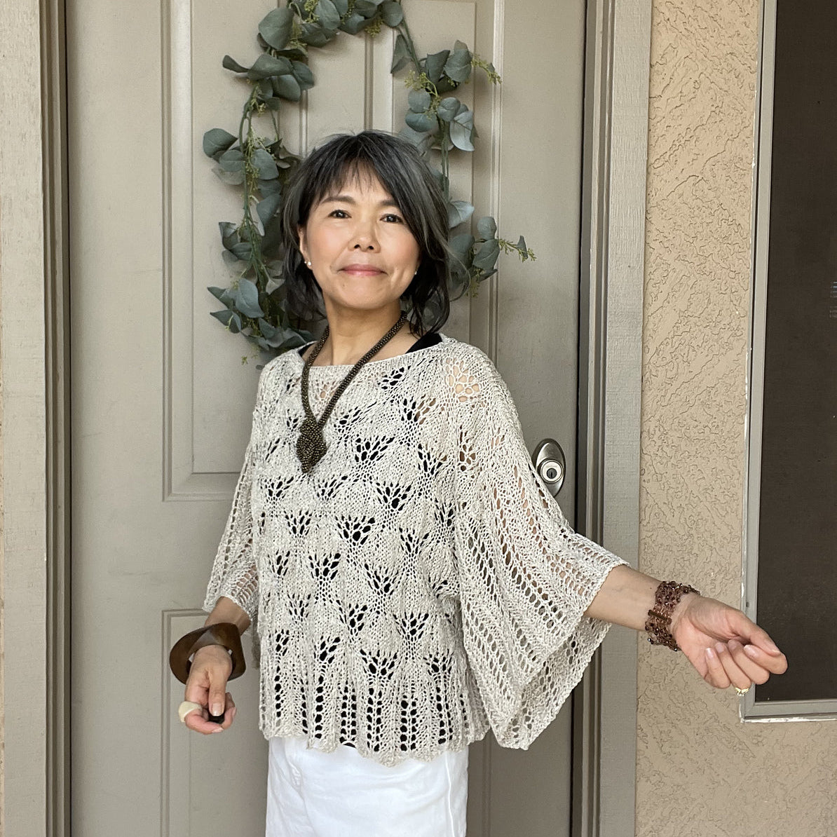 Woman standing in front of a door with a wreath, wearing a white lace top and white pants.